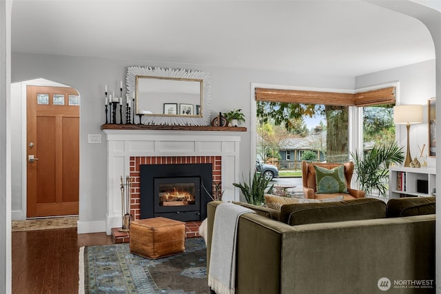 living room with arched walkways, a brick fireplace, baseboards, and wood finished floors