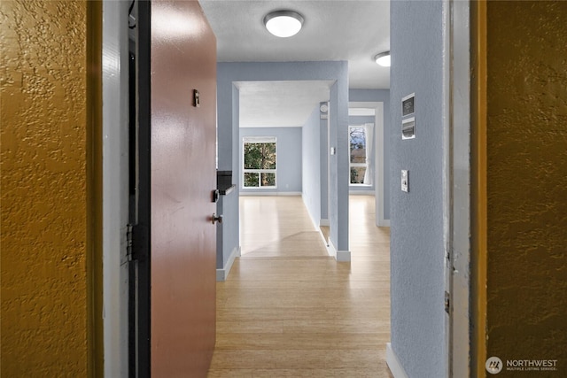 corridor with light wood-type flooring, a textured wall, and baseboards