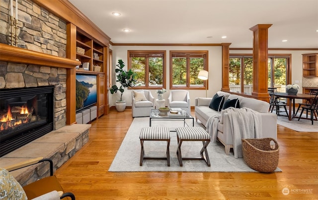 living room featuring light wood-type flooring, a stone fireplace, crown molding, and decorative columns