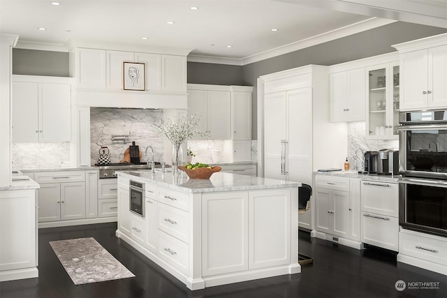 kitchen featuring white cabinetry, tasteful backsplash, light stone countertops, an island with sink, and stainless steel double oven