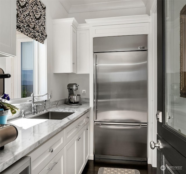 kitchen featuring built in fridge, white cabinetry, sink, ornamental molding, and light stone counters