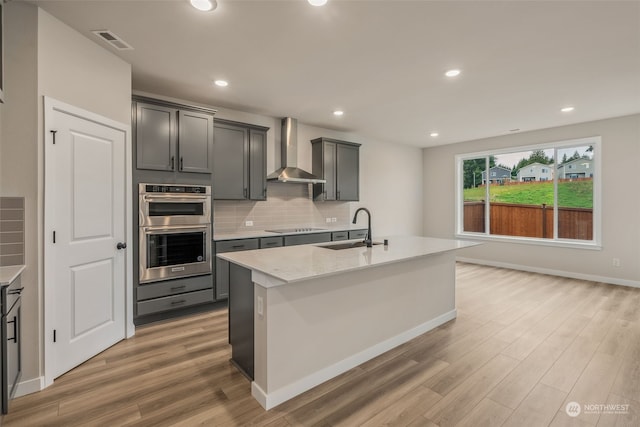 kitchen featuring double oven, wall chimney exhaust hood, sink, and a center island with sink