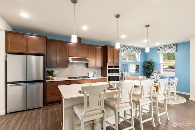 kitchen featuring hanging light fixtures, appliances with stainless steel finishes, dark hardwood / wood-style flooring, a kitchen island with sink, and backsplash