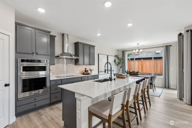 kitchen featuring double oven, wall chimney exhaust hood, sink, black electric stovetop, and an island with sink