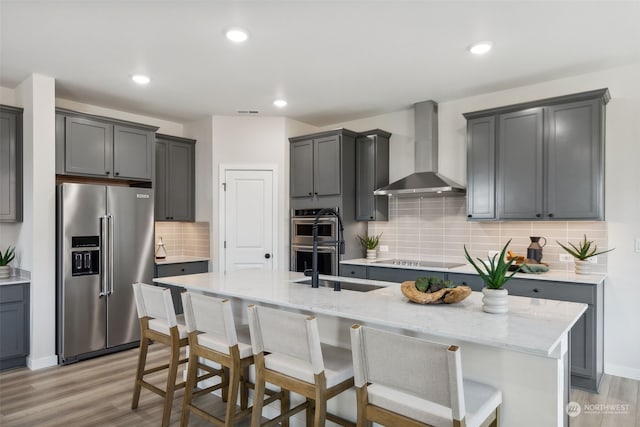 kitchen featuring high quality fridge, a kitchen island with sink, double wall oven, wall chimney range hood, and a breakfast bar area