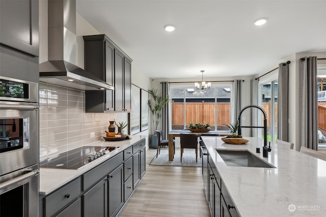 kitchen featuring wall chimney exhaust hood, sink, decorative light fixtures, black electric stovetop, and light stone counters