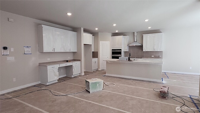 kitchen with wall chimney range hood, backsplash, a center island with sink, baseboards, and white cabinetry
