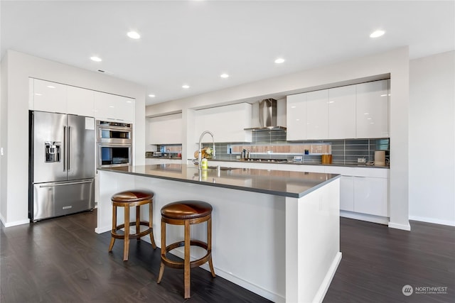 kitchen featuring wall chimney range hood, white cabinets, appliances with stainless steel finishes, and a center island with sink