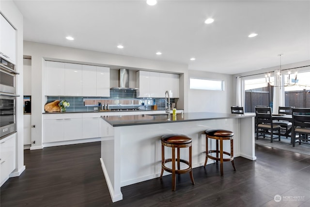 kitchen featuring wall chimney exhaust hood, white cabinets, a kitchen island with sink, and sink