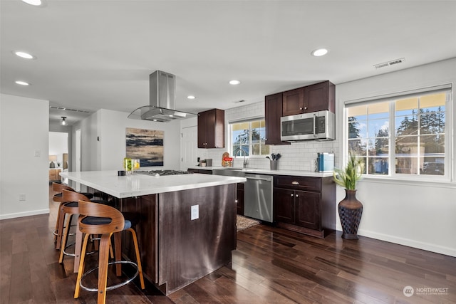 kitchen with dark wood-type flooring, tasteful backsplash, island exhaust hood, a center island, and stainless steel appliances