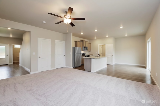 kitchen featuring stainless steel appliances, an island with sink, ceiling fan, light stone counters, and dark carpet