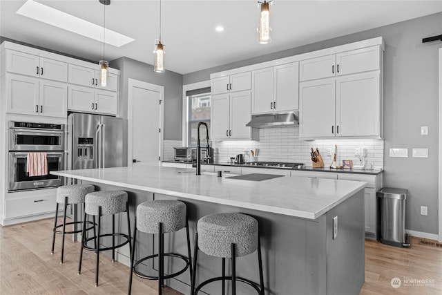 kitchen with appliances with stainless steel finishes, white cabinetry, backsplash, hanging light fixtures, and a kitchen island with sink