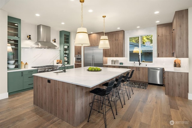 kitchen with appliances with stainless steel finishes, a kitchen island with sink, green cabinets, wall chimney range hood, and pendant lighting