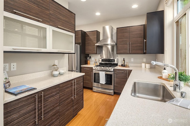 kitchen featuring dark brown cabinetry, sink, light wood-type flooring, appliances with stainless steel finishes, and wall chimney range hood