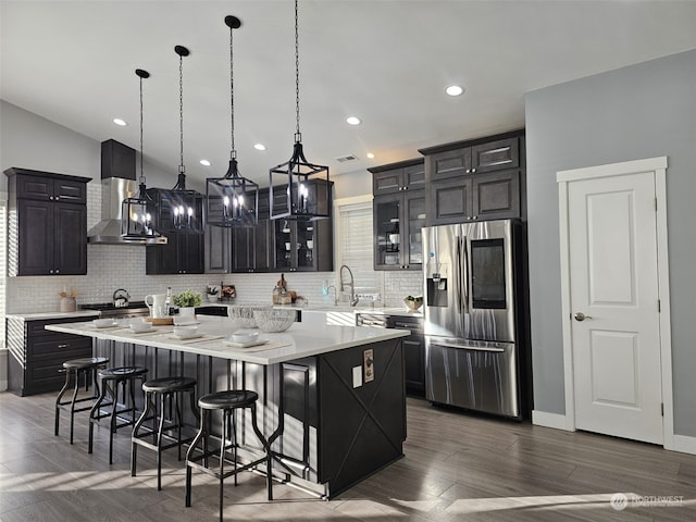 kitchen featuring a large island, a breakfast bar area, stainless steel refrigerator with ice dispenser, decorative light fixtures, and wall chimney exhaust hood