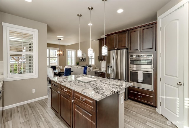 kitchen with hanging light fixtures, stainless steel appliances, dark brown cabinetry, light stone counters, and a kitchen island