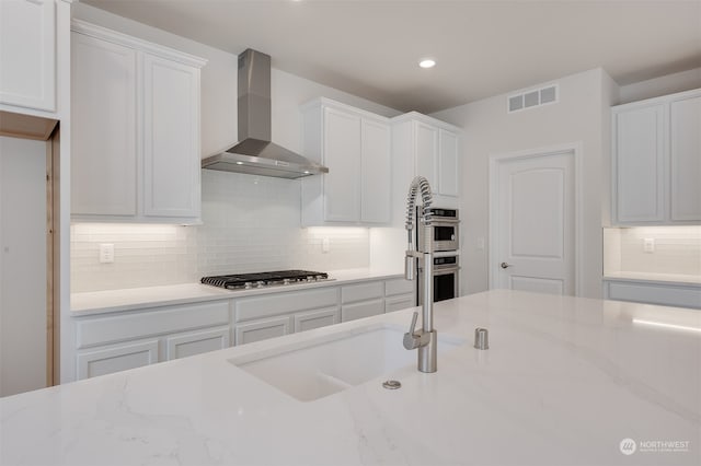 kitchen with white cabinetry, decorative backsplash, stainless steel appliances, light stone countertops, and wall chimney range hood