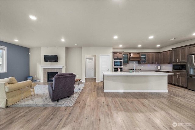 kitchen featuring premium range hood, dark brown cabinetry, light hardwood / wood-style flooring, appliances with stainless steel finishes, and an island with sink