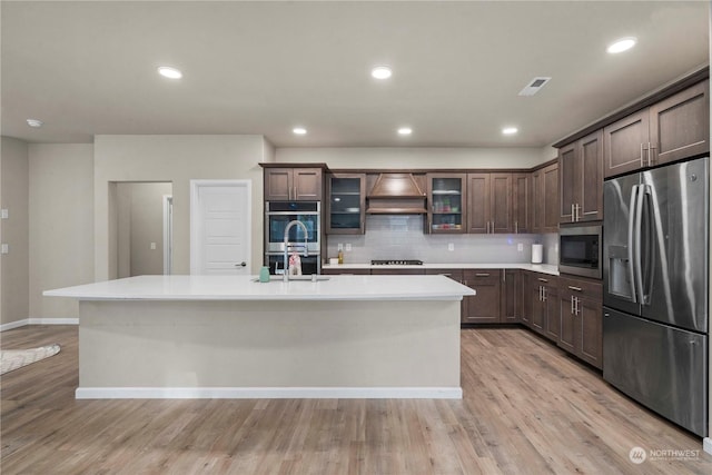 kitchen with light hardwood / wood-style flooring, appliances with stainless steel finishes, dark brown cabinets, a center island with sink, and custom exhaust hood