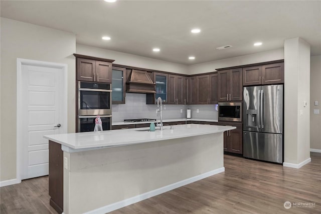 kitchen featuring dark brown cabinets, stainless steel appliances, custom range hood, and a center island with sink