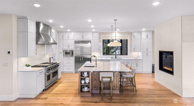 kitchen with built in appliances, an island with sink, a kitchen bar, wall chimney exhaust hood, and light wood-type flooring