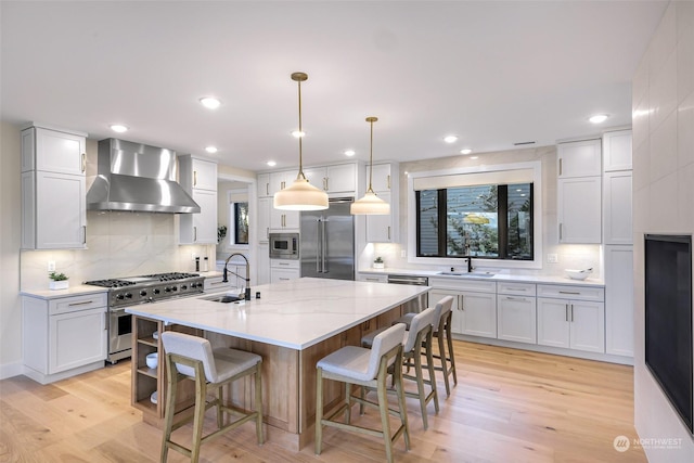 kitchen with wall chimney exhaust hood, sink, built in appliances, an island with sink, and white cabinets