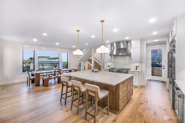 kitchen with pendant lighting, white cabinetry, an island with sink, light stone counters, and wall chimney range hood