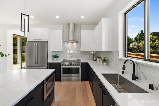 kitchen featuring wall chimney exhaust hood, sink, white cabinetry, premium appliances, and pendant lighting