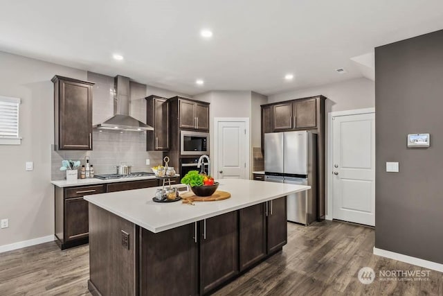 kitchen with stainless steel appliances, dark brown cabinets, a center island with sink, and wall chimney exhaust hood