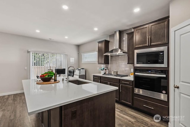 kitchen with wall chimney range hood, sink, appliances with stainless steel finishes, backsplash, and a center island with sink