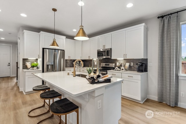 kitchen with white cabinetry, appliances with stainless steel finishes, a kitchen island with sink, pendant lighting, and light hardwood / wood-style floors