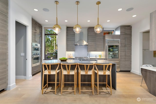 kitchen with decorative light fixtures, a kitchen island with sink, light hardwood / wood-style flooring, and wall chimney range hood