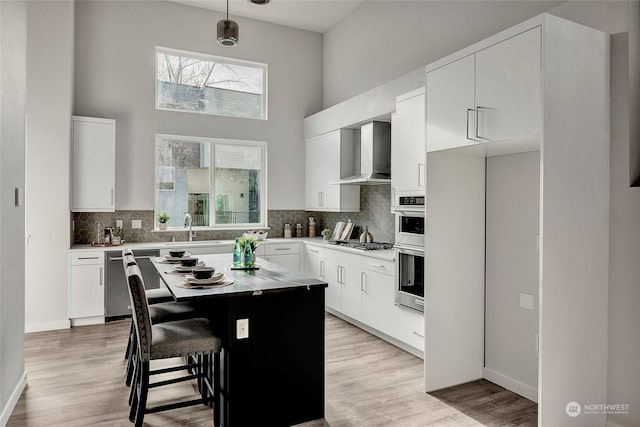 kitchen featuring wall chimney exhaust hood, a center island, hanging light fixtures, appliances with stainless steel finishes, and white cabinets