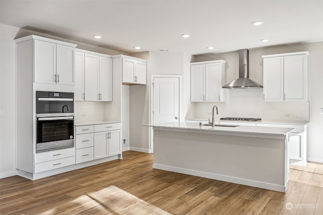 kitchen featuring wall chimney range hood, sink, light hardwood / wood-style flooring, white cabinets, and a center island with sink