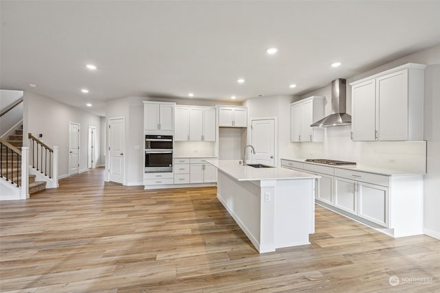 kitchen featuring wall chimney exhaust hood, sink, white cabinetry, stainless steel appliances, and a kitchen island with sink