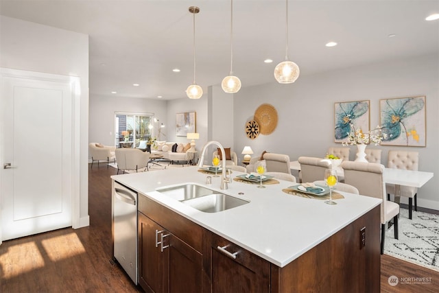 kitchen with an island with sink, sink, hanging light fixtures, stainless steel dishwasher, and dark wood-type flooring