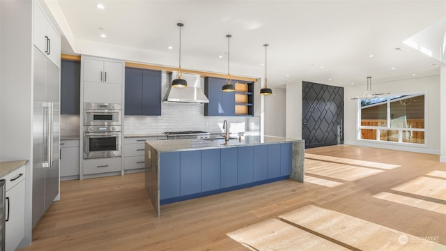 kitchen with wall chimney range hood, decorative light fixtures, an island with sink, and stainless steel double oven