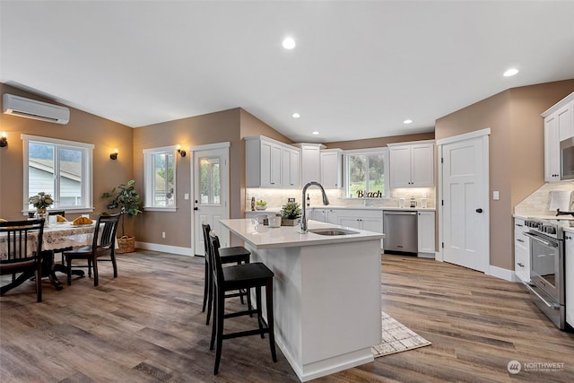 kitchen with sink, appliances with stainless steel finishes, white cabinetry, a wall mounted AC, and a center island with sink