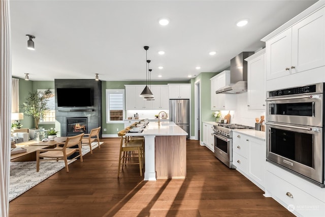 kitchen featuring decorative light fixtures, high quality appliances, white cabinets, a kitchen island with sink, and wall chimney range hood