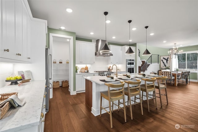 kitchen with white cabinetry, a kitchen island with sink, wall chimney range hood, and decorative light fixtures