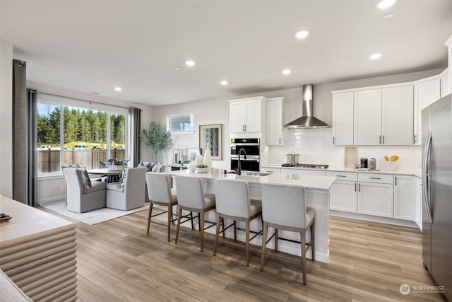 kitchen with white cabinets, stainless steel appliances, a center island with sink, and wall chimney range hood