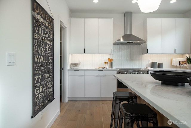 kitchen with wall chimney range hood, tasteful backsplash, light stone countertops, light hardwood / wood-style floors, and white cabinets