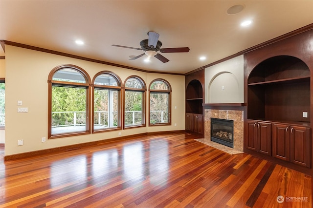 unfurnished living room with built in shelves, ornamental molding, a tiled fireplace, and hardwood / wood-style flooring