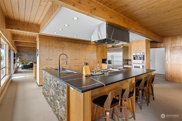 kitchen with beam ceiling, stainless steel appliances, island range hood, light carpet, and wood walls