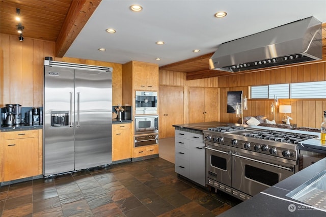 kitchen featuring wood walls, built in appliances, beam ceiling, and wall chimney range hood