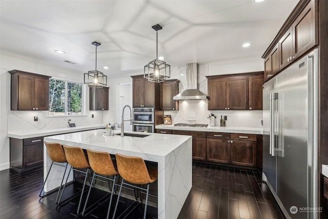 kitchen featuring pendant lighting, sink, stainless steel appliances, a center island with sink, and wall chimney exhaust hood