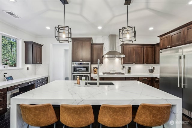 kitchen with pendant lighting, wall chimney range hood, stainless steel appliances, and an island with sink