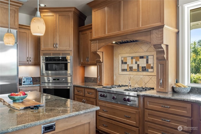 kitchen with appliances with stainless steel finishes, tasteful backsplash, dark stone countertops, hanging light fixtures, and custom exhaust hood