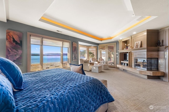 bedroom featuring carpet flooring, access to exterior, a tray ceiling, a premium fireplace, and a water view