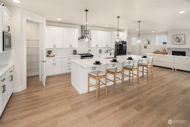 kitchen with built in microwave, wall chimney exhaust hood, a kitchen island with sink, and white cabinets